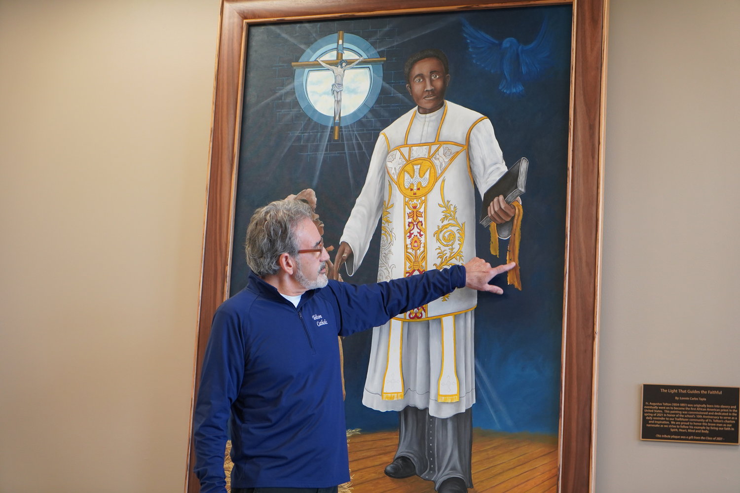 Lonnie Carlos Tapia, who teaches art, photography and design at Fr. Tolton Regional Catholic High School in Columbia, draws attention to symbols he incorporated into the new painting of Venerable Fr. Augustus Tolton commissioned for the school’s 10th anniversary, during a May 14 unveiling and dedication ceremony. The school’s Class of 2021 donated the plaque beside the painting, which hangs in a busy area next to the chapel of the school building.