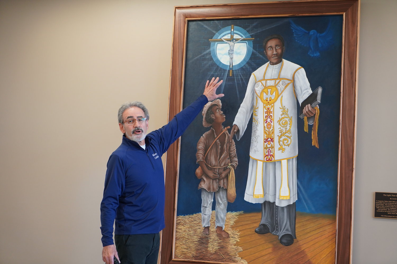 Lonnie Carlos Tapia, who teaches art, photography and design at Fr. Tolton Regional Catholic High School in Columbia, draws attention to symbols he incorporated into the new painting of Venerable Fr. Augustus Tolton commissioned for the school’s 10th anniversary, during a May 14 unveiling and dedication ceremony. The school’s Class of 2021 donated the plaque beside the painting, which hangs in a busy area next to the chapel of the school building.