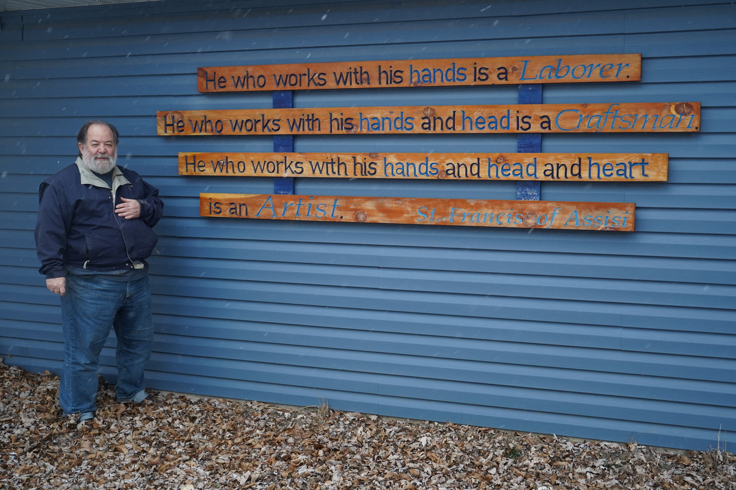 Fr. Means stands outside his workshop, next to a sign containing a quote from St. Francis Assisi.